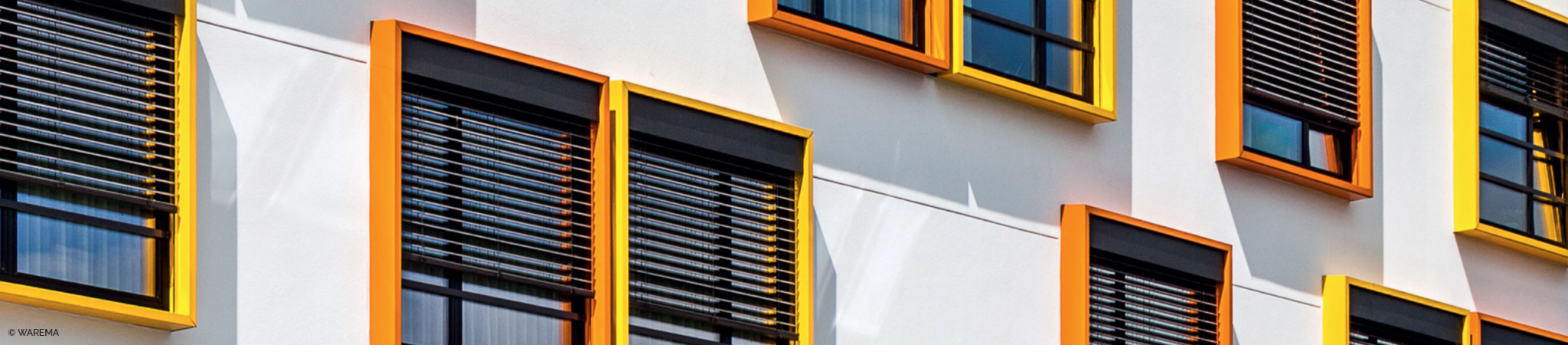 Fassadenansicht mit mehreren Fenstern in orangefarbenen Rahmen und heruntergelassenen Außenjalousien als Sonnenschutz.
