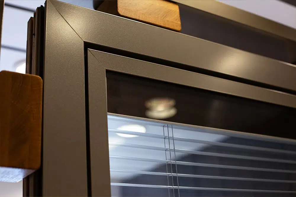 Nahaufnahme eines anthrazitfarbenen Metallfensters mit eingebauten Jalousien und sichtbaren Holzelementen sowie Lichtreflexion im Glas