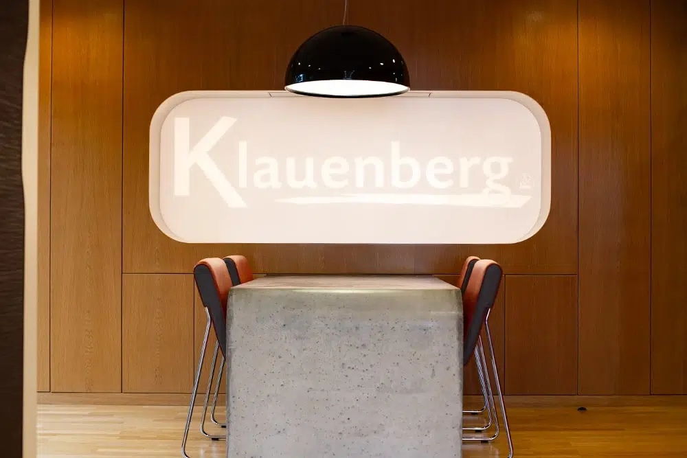 Moderner Besprechungsbereich mit Betontisch, vier Stühlen, Hängeleuchte und beleuchtetem Wandfenster mit dem Schriftzug Klauenberg