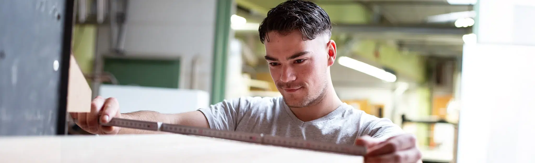 Person misst eine Holzplatte mit einem Zollstock in einer Werkstatt.