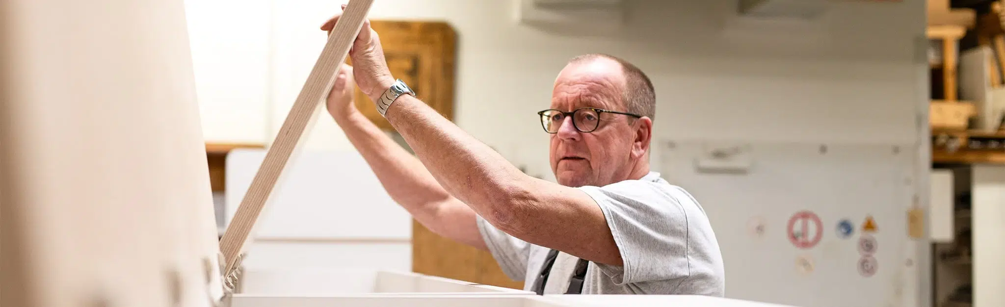 Handwerker mit Brille hebt in einer hellen Werkstatt eine Holzplatte.