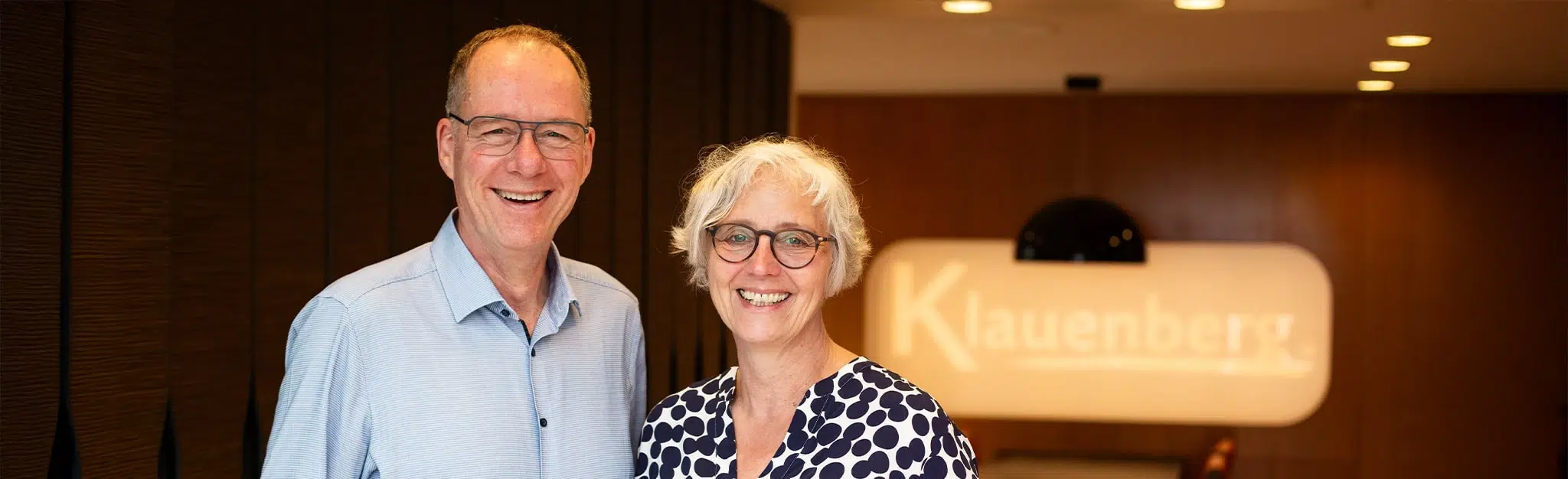 Startseiten-Headerbild mit zwei lächelnden Personen (Mann und Frau) im Empfangsbereich vor einem unscharfen Firmenlogo.