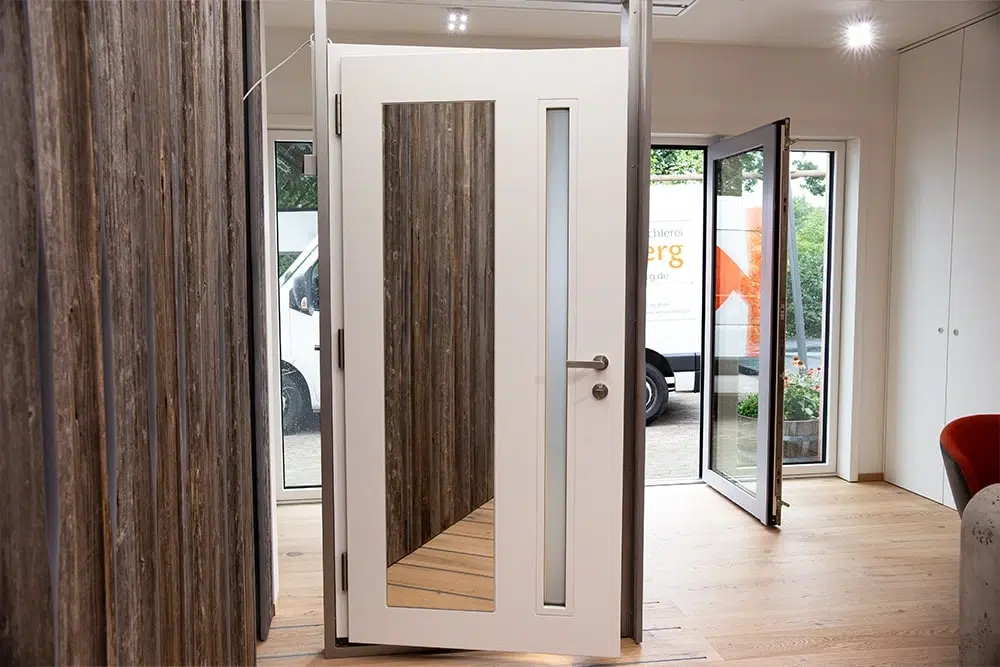 Ausstellungsraum mit offenstehender weißer Innentür mit schmalem Glasfenster vor Holzverkleidung und geöffneter Außentür zum Hof.