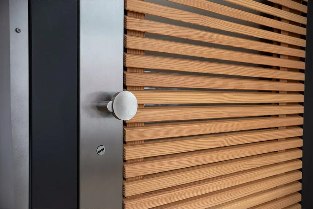 Detailaufnahme einer modernen Tür mit horizontalen Holzlamellen, Metallrahmen und rundem Edelstahlknauf.