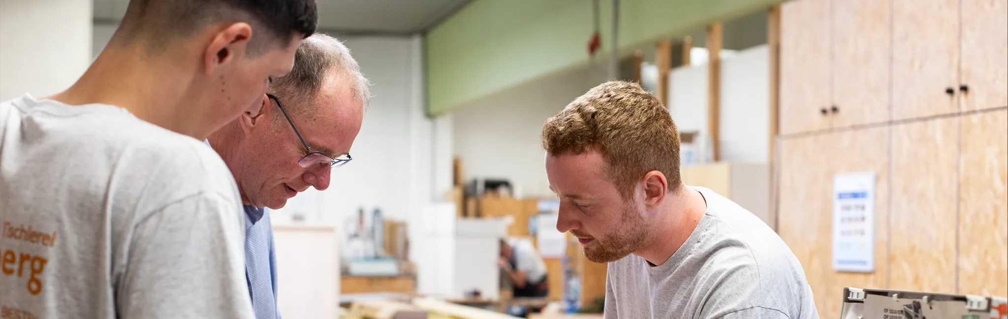 Älterer Ausbilder erklärt zwei Auszubildenden in einer Werkstatt eine praktische handwerkliche Aufgabe.