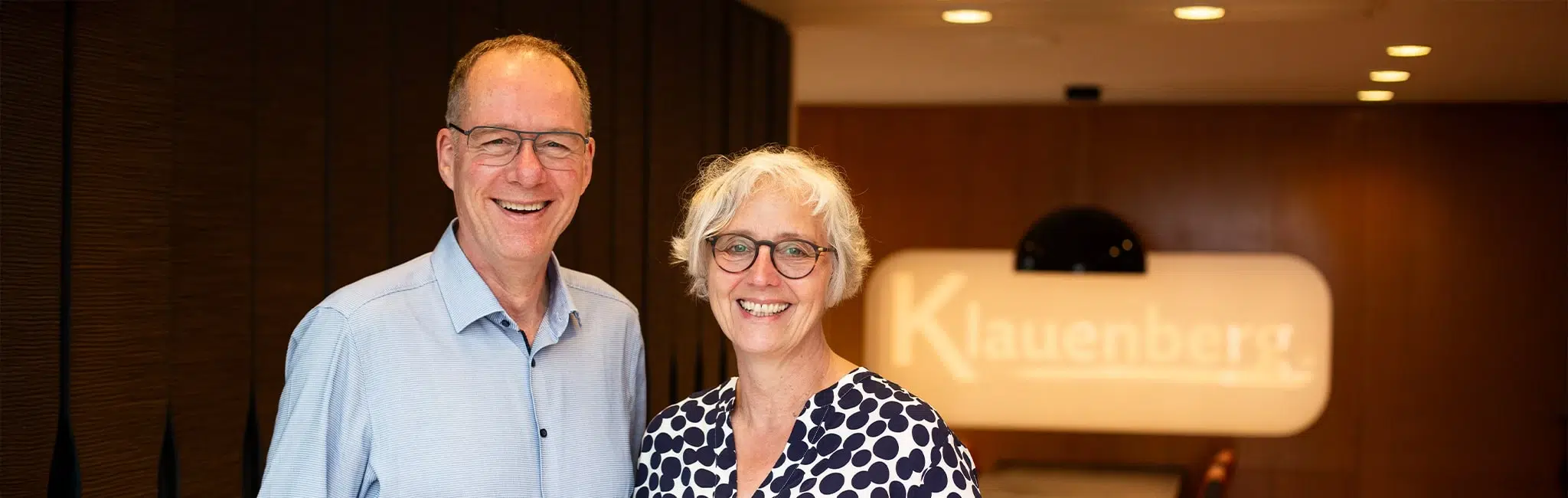 Headerbild der Kontaktseite: Zwei lächelnde Personen (Mann und Frau) in einem Empfangsbereich, im Hintergrund unscharfes Leuchtschild mit Firmenlogo.