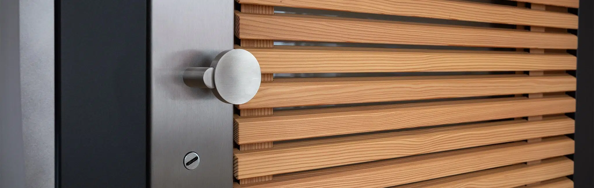 Nahaufnahme eines runden Edelstahl-Türgriffs an einer Holzlamellen-Tür mit dunklem Metallrahmen.