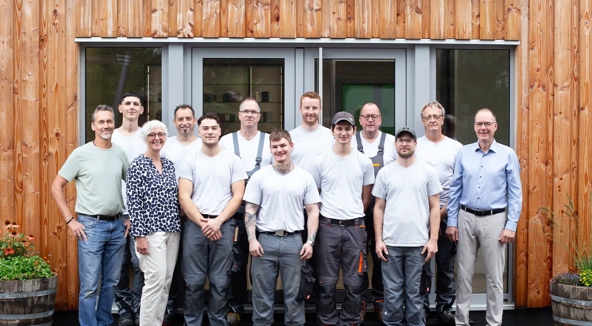 Teamfoto eines Handwerksbetriebs: Mitarbeitende in Arbeitskleidung und zwei Personen in Alltagskleidung stehen vor einer Holzfassade mit Glastür.